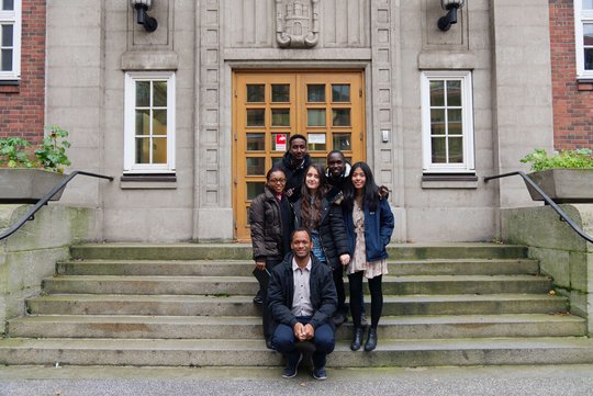 Eine Gruppe von 6 jungen Forschern, die als fröhliche Gruppe auf einer steinernden Treppe vor einem Antiken Gebäude stehen.