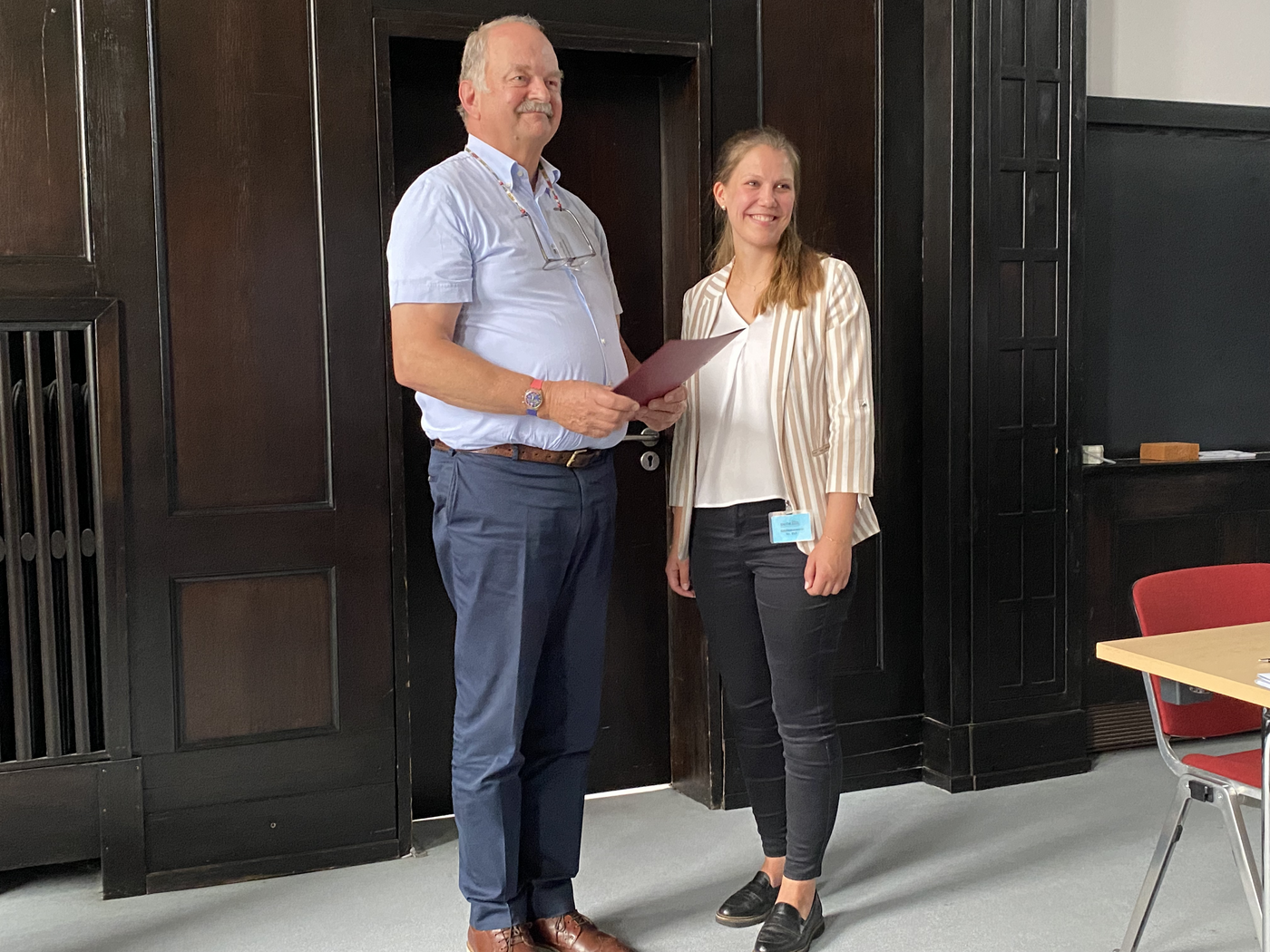 A board member of the Association of Friends of the Tropical Institute presents Marie Groneberg with a certificate at the award ceremony in the lecture theatre of the Bernhard Nocht Institute. In the background you can see a wall panelled in dark brown.