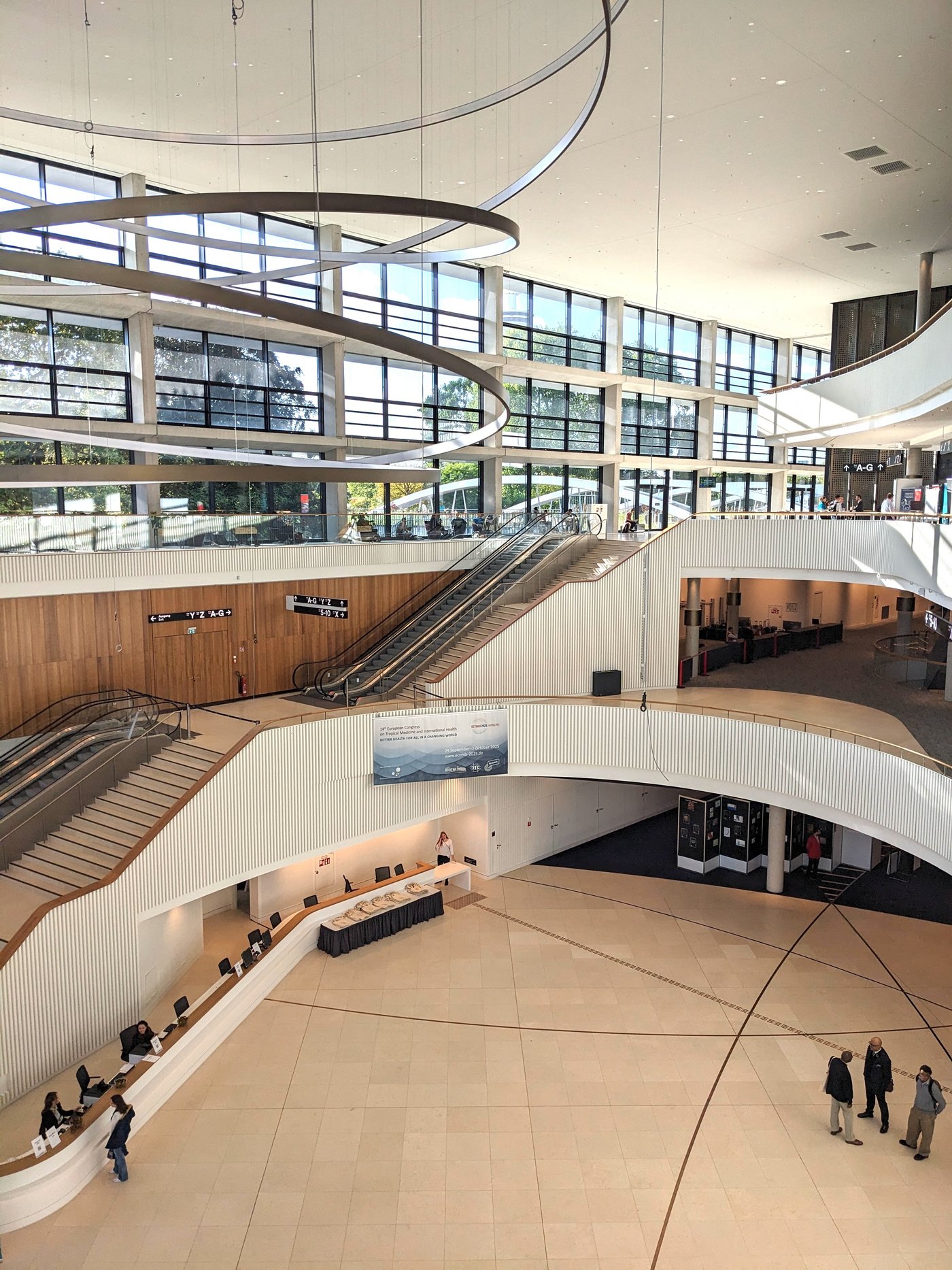 The picture shows the large reception hall of the CCH Hamburg. The staircase is very modern in beige and wood colors. A large lighting object made of steel circles hangs from the roof. The photo was taken from an upper floor.