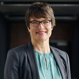 Julia Rauner: a woman short hair and glasses. She wears a petrol-colored blouse and a gray blazer.