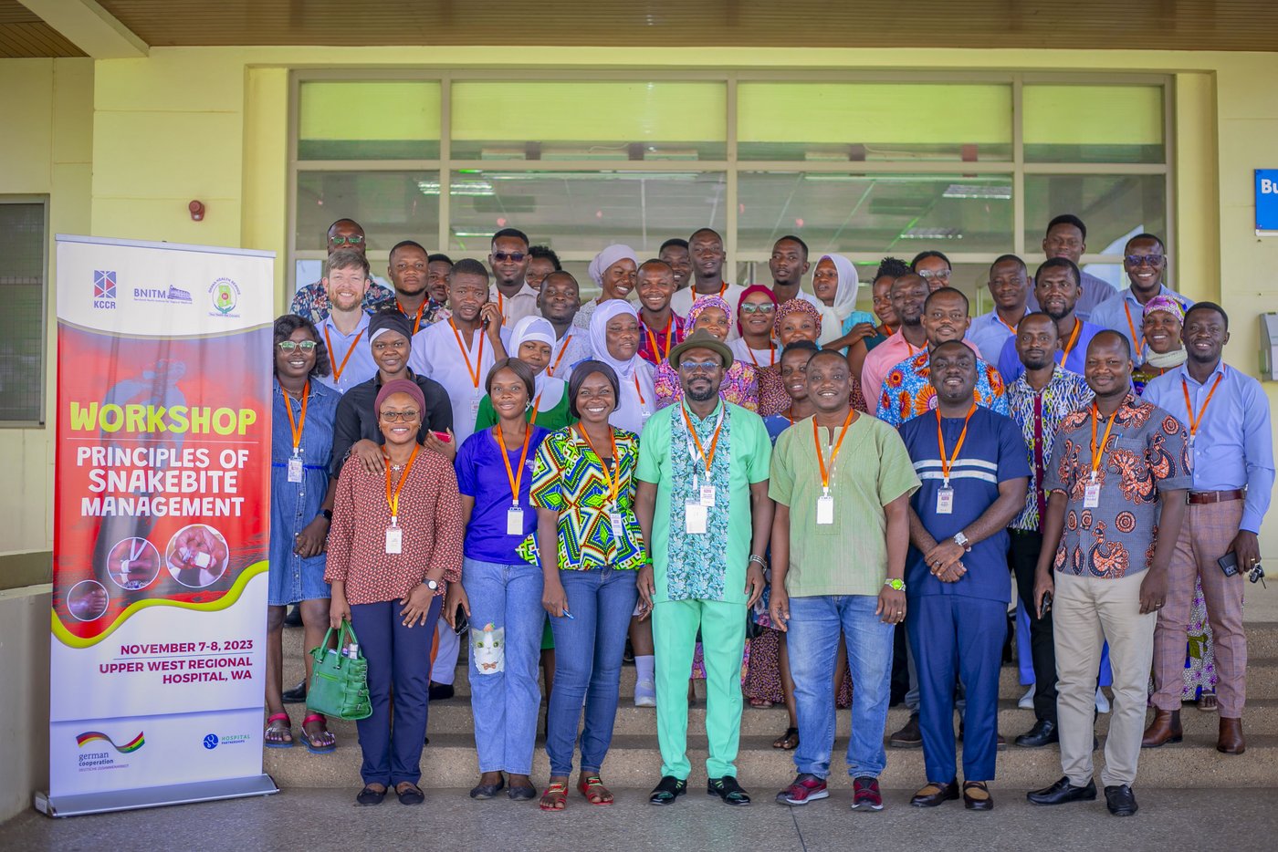 Das Bild zeigt eine Gruppe von Menschen, die vor dem Eingang eines Krankenhauses stehen, neben einem Banner mit der Aufschrift "Workshop - Principles of Snakebite Mangement". Alle schauen freundlich in die Kamera.