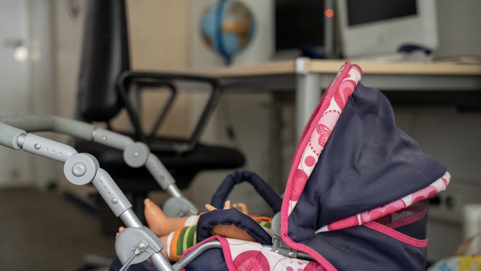 Das Familienzimmer Das Foto zeigt einen Spielzeug-Kinderwagen, im Hintergrund ein Schreibtisch-Arbeitsplatz.