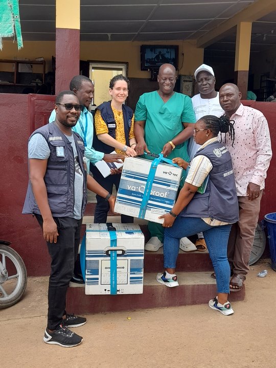 Seven people in colourful clothes, three with WHO vests, are standing on the door step of a building in Guinea. In front of them are two big white and blue boxes that have just been delivered.