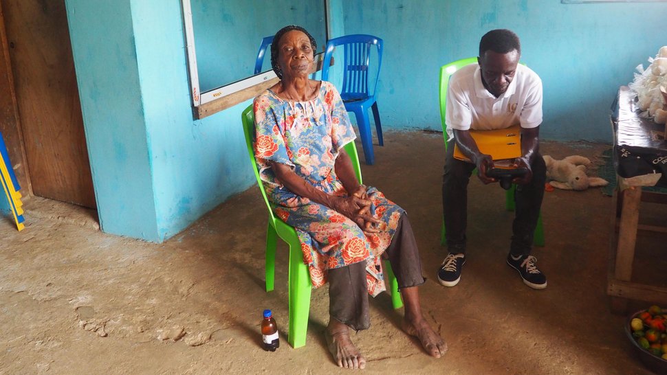 An elderly barefoot woman and a younger man are sitting in a waiting room with mint-coloured walls in Gabon, waiting on bright green plastic chairs.