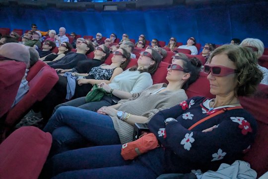 The picture shows visitors to the star show inside the planetarium. They are sitting back in comfortable armchairs and wearing 3D glasses. The lights have already been dimmed for the show.