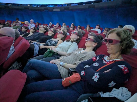 The picture shows visitors to the star show inside the planetarium. They are sitting back in comfortable armchairs and wearing 3D glasses. The lights have already been dimmed for the show.