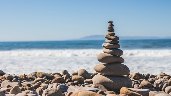 Ein Strand mit großen Kieselsteinen, aus denen mit mehreren, kleiner werdenden Kieselsteinen ein kleiner Turm gebaut wurde.