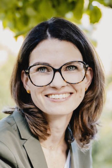 Porträtfoto von Prof. Cornelia Betsch. Eine Frau mit schulterlangen braunen Haaren und Brille sitzt sehr freundlich lächelnd unter einem Baum. Sie trägt einen olivgrünen-grauen Anzug mit weißem Shirt darunter.