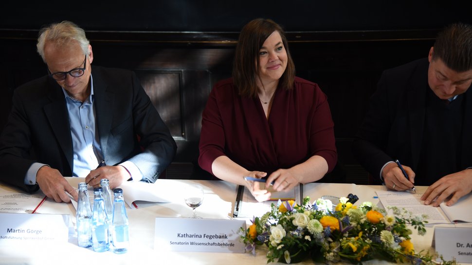 Das Bild zeigt von links nach rechts Martin Görge (Sprinkenhof), Katharina Fegebank (Wissenschaftsbehörde) und Dr. Andreas Dressel (Finanzbehörde) bei der Unterzeichnung des Letter of Intend. Frau Fegebank blickt lächelnd hoch, während die anderen noch schreiben. Vor ihr steht ein buntes Blumengesteck.