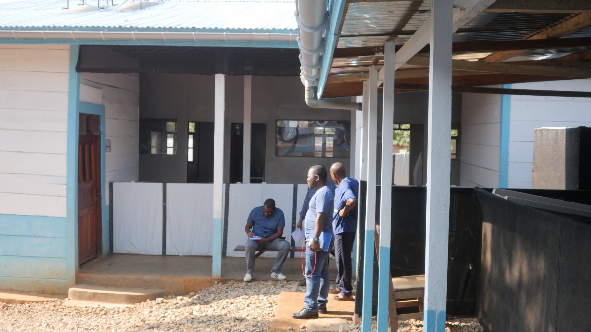 Several men are standing outside at a veranda, one is sitting on a bench writing something in a notepad.