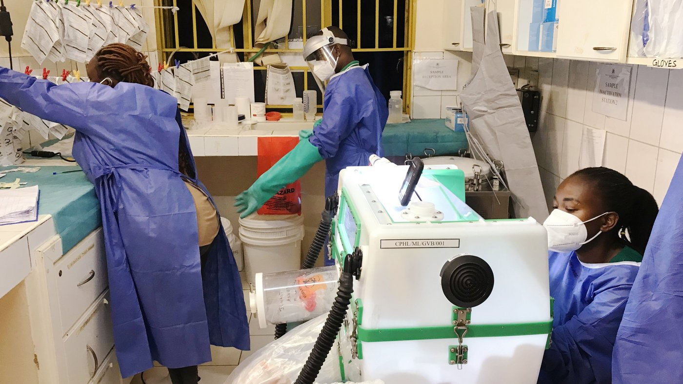 The scene takes place in the evening. Three scientific staff members are in a laboratory room processing incoming suspected Ebola virus samples. One of them is wearing full personal protective equipment, including a face shield and long green rubber gloves, to receive the suspect samples through the lab window above a work bench. A second person hangs decontaminated lab requisition forms on a line to dry. The third person sits in front of a glovebox and prepares to inactivate the suspect samples.
