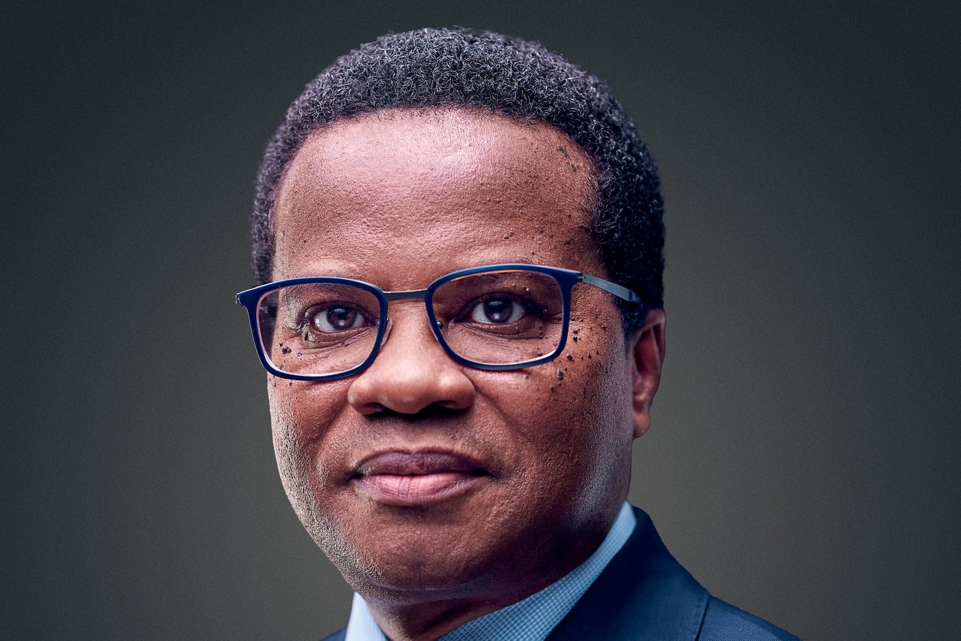 The picture shows a self-confident middle-aged African scientist in a blue suit with a light blue tie and black-rimmed glasses.