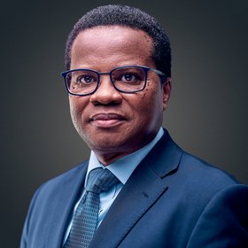 The picture shows a self-confident middle-aged African scientist in a blue suit and light blue tie plus black-rimmed glasses.