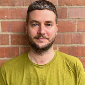 PhD Dominik Vogel: A researcher looks into the camera. He wears short brown-grey hair, a three-day beard and a curry-coloured shirt.