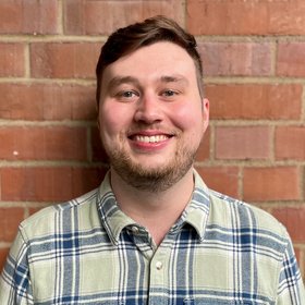 PhD Harry Williams: A researcher smiles at the camera. He wears short brown hair, a short beard and a checked shirt.