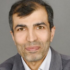A close-up portrait of a middle-aged man wearing a gray blazer over a white shirt, with short dark hair and a slight beard, smiling against a gray background.