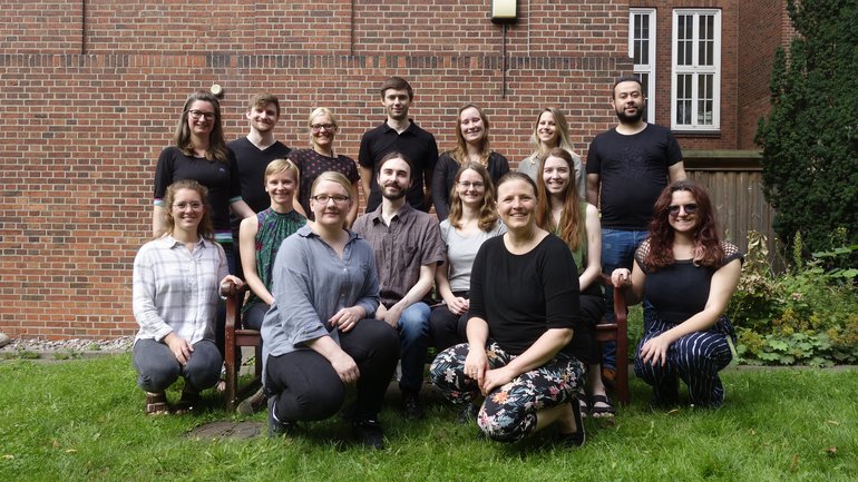 Foto der Laborgruppe Oestereich. Die Mitglieder der Leibniz Junior Laborgruppe Oestereich sind hier dargestellt. Einige Personen stehen, andere sitzen auf oder knien vor einer Bank, alle mit einem Lächeln im Gesicht.