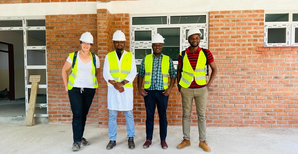 The picture shows four people wearing high-visibility vests and safety helmets in front of a building. They are all smiling at the camera.