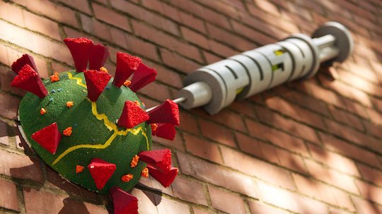 Photograph of wall art on a brick wall in sunlight. It shows a coronavirus with a syringe with the words "Push".