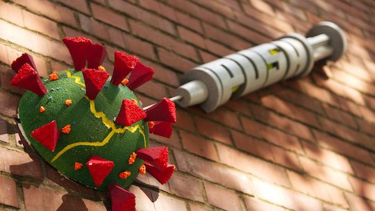 Künstlerische Skulptur des SARS-Cov-2-Virus und einer Spritze mit Link zur Coronadiagnostik und -beratung