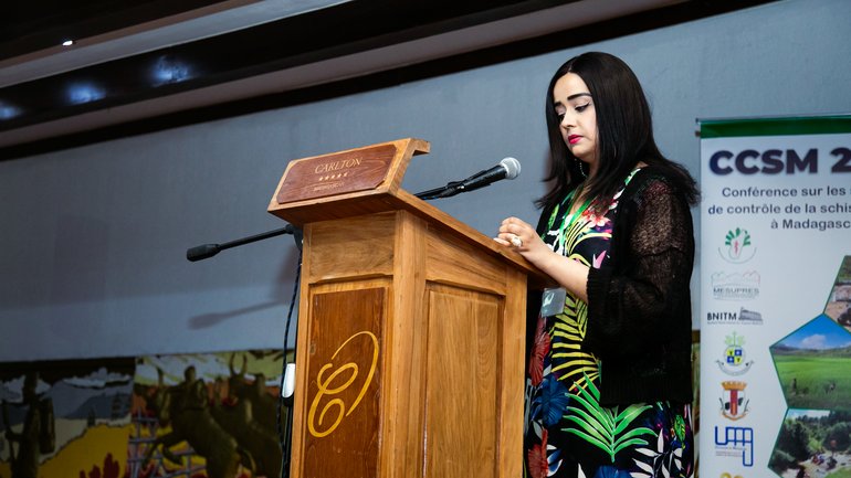 A woman presenting at the CCSM 2024 conference at a wooden podium, wearing a colorful dress, with a CCSM 2024 conference banner in the background.