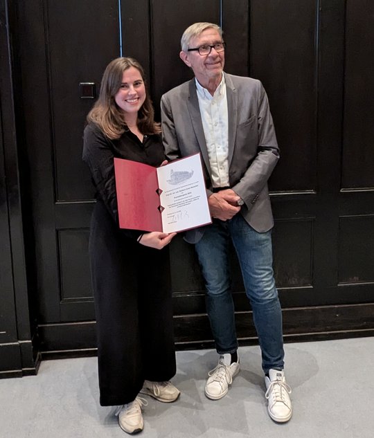 A board member of the Association of Friends of the Tropical Institute presents Barbara Honecker with a certificate at the award ceremony in the lecture theatre of the Bernhard Nocht Institute. In the background you can see a wall panelled in dark brown
