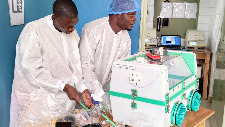 The photo shows two men in white lab coats setting up a glove box.