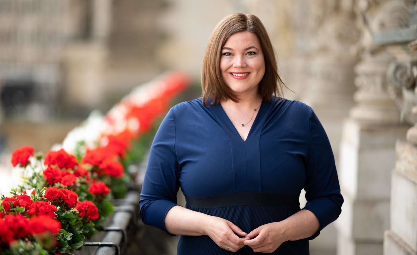 Katharina Fegebank: a politician on a balcony. On the left are red flowers, on the right the light-coloured walls of Hamburg City Hall. Katarina Fegebank wears shoulder-length brown hair and a blue dress. The fingertips of her two hands touch in front of the centre of her body.