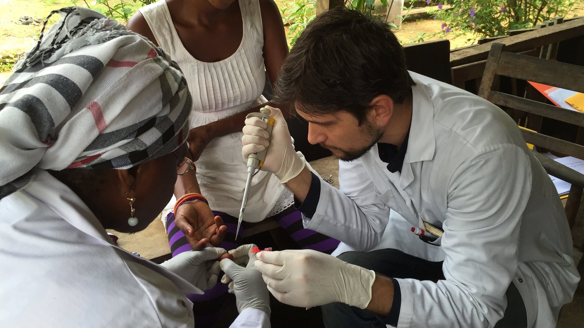 Mischlinger im Feldeinsatz: Zu sehen ist Dr. Mischlinger, der mit einer Pipette Blut von dem Finger einer Patienten in ein Eppie pippetiert. Eine Helferin hält den Finger der Patientin fest.