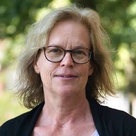 Sabine Fehrmann: eine Frau mit halblangen Haaren und Brille. Sie trägt ein fliederfarbenes T-Shirt mit schwarzer Jacke. Im Hintergrund sind unscharf Blätter und Sonne zu sehen.