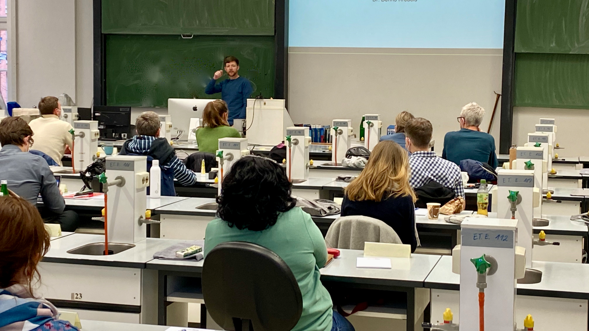 The photo shows the course room with course participants from behind. The green board can be seen in front, a lecturer is standing in front of it.
