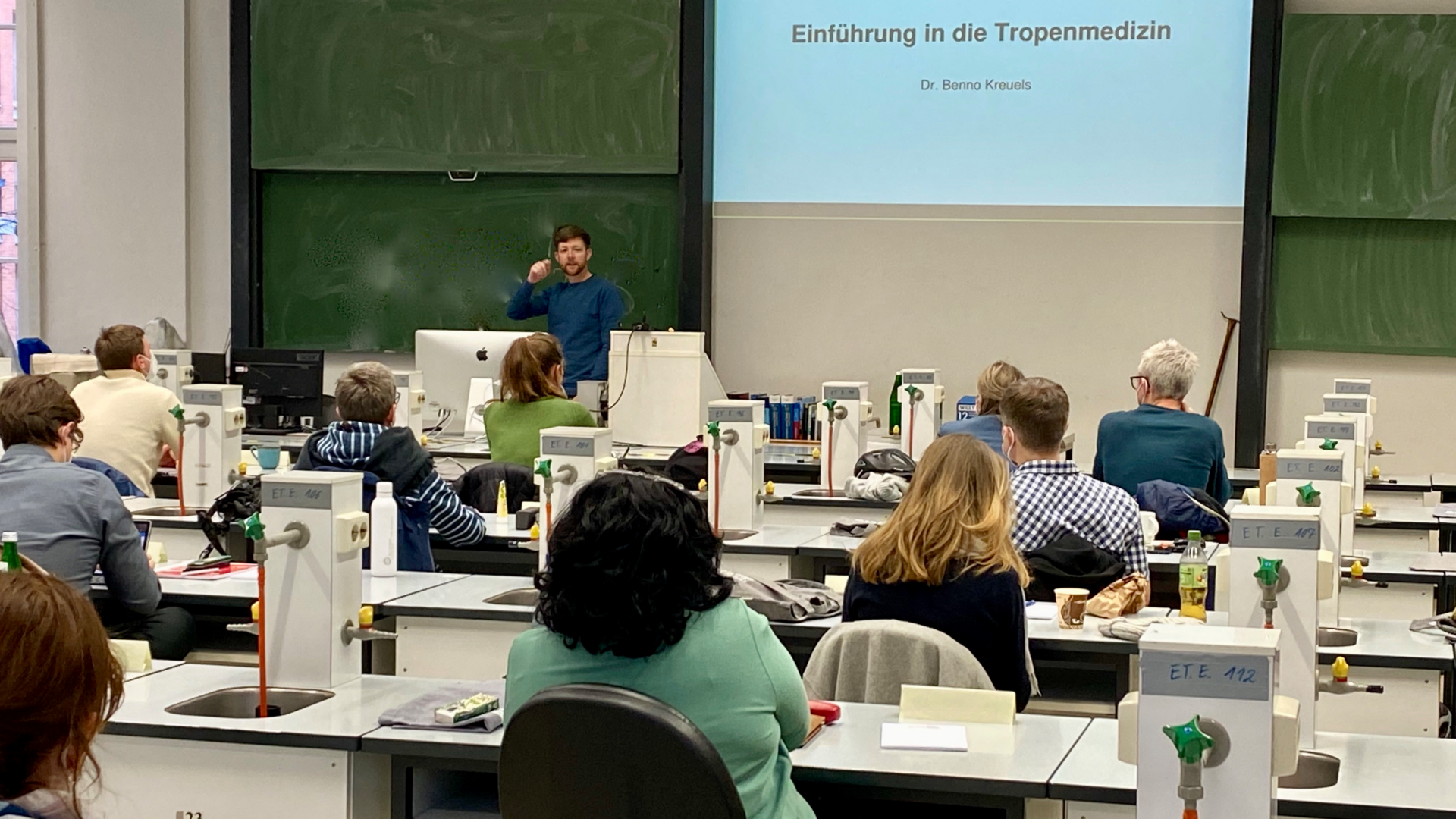 Das Foto zeigt den Kursussaal mit Kursteilnehmenden von hinten. Vorn ist die grüne Tafel zu sehen, davor steht ein Dozent.