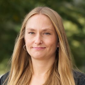 Jule Hinzmann Jule Hinzmann: eine lächelnde Frau mit langen blonden Haaren und Ohrringen. Sie trägt ein schwarzes T-Shirt und eine Jacke und steht vor einem unscharfen Blätterhintergrund.