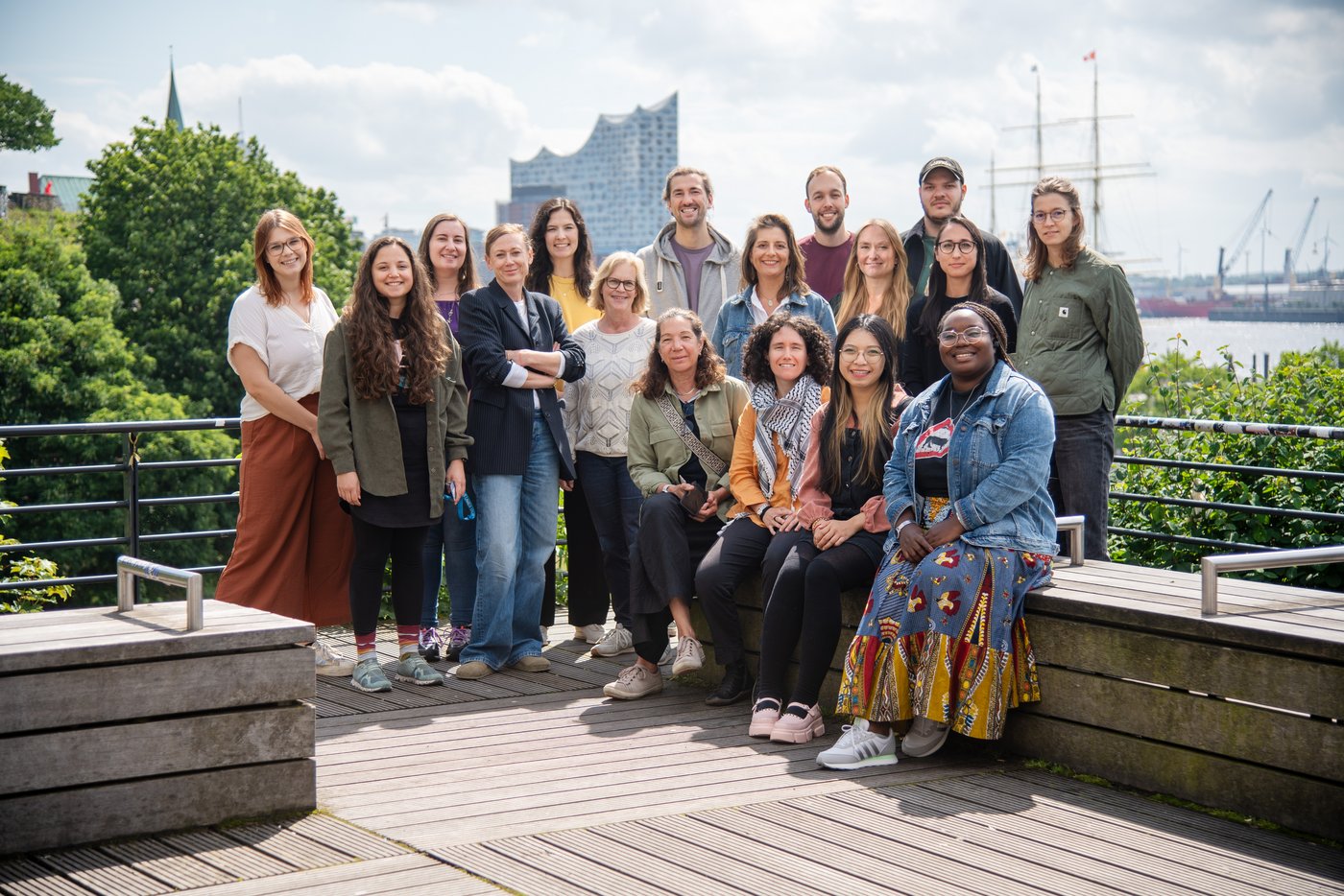 Team of the Research Group Duraffour-Pahlmann - Outbreak Preparedness and Response The picture shows a team of 17 people sitting on a bench or standing behind it on a sunny day. In the background you can see the building of the Hamburg Elbphilharmonie and the masts of a sailing ship.
