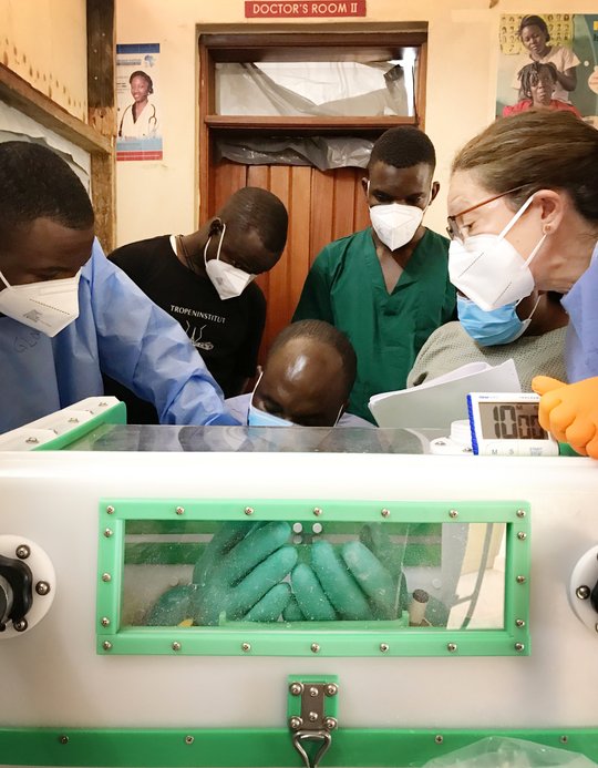 Drei lokale Labormitarbeitende, ein Ausbilder, der eine persönliche Schutzausrüstung (blauer Kittel und orangefarbene Handschuhe) trägt, und zwei Auszubildende, die ebenfalls eine ähnliche persönliche Schutzausrüstung tragen, stehen um eine Glovebox herum. Ein Auszubildender hat seine Hände in der Glovebox. Er wechselt sicher die Handschuhe, die mit der Glovebox verbunden sind, indem er die alten Handschuhe durch neue ersetzt. Dabei wird er von einem zweiten Auszubildenden unterstützt. Der Ausbilder beaufsichtigt und leitet den Vorgang des Handschuhwechsels an. Die drei anderen Teilnehmer:innen beobachten den Vorgang.