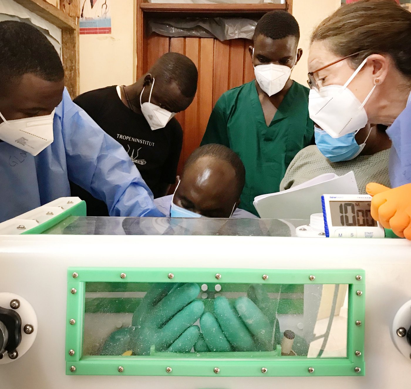 Three local laboratory employees, an instructor wearing personal protective equipment (blue smock and orange gloves), and two trainees also wearing similar personal protective equipment are standing around a glovebox. One trainee has his hands in the glovebox. He safely changes the gloves connected to the glovebox by replacing the old gloves with new ones. He is assisted by a second trainee. The instructor supervises and guides the glove changing process. The other three trainees observe the process.