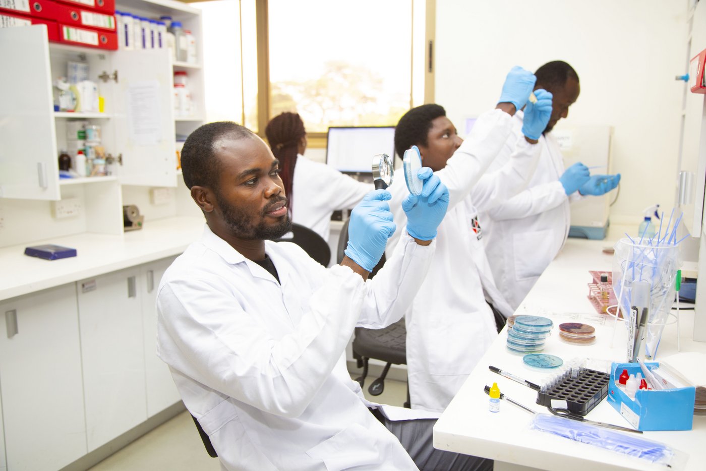 The picture shows three African researchers and one African woman researcher in white coats and light blue gloves working in the laboratory.