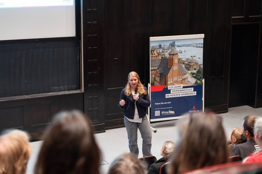 Auf dem Foto sieht man Lisa Oestereich im Hörsaal neben einem Plakat der 125-Jahrfeier des BNITM stehen und einen Vortrag halten.
