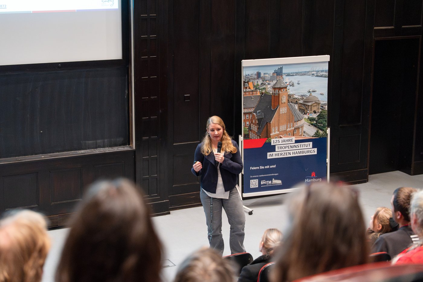 Lisa Oestereich Auf dem Foto sieht man Lisa Oestereich im Hörsaal neben einem Plakat der 125-Jahrfeier des BNITM stehen und einen Vortrag halten.