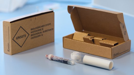 An example of a correct sample shipment is shown: A small brown cardboard box with the inscription UN3373. A milky plastic tube lies in front of it, lying open. Half of a blood tube is visible from the opening. The lid of the milky plastic tube lies in front of it.