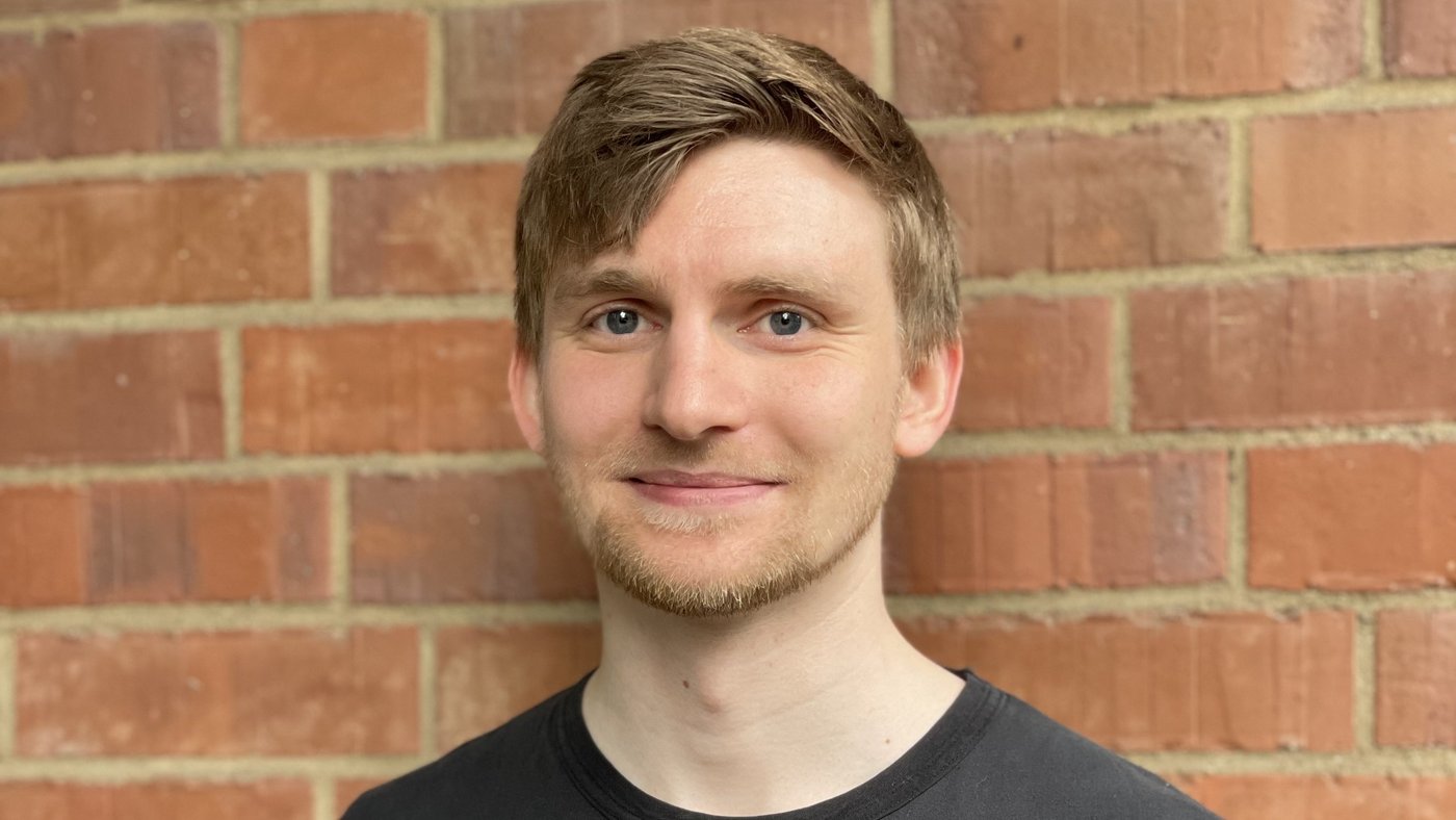 The photo shows a young junior researcher with short dark blond hair and a slight beard in a black T-shirt standing in front of a red brick wall and smiling.