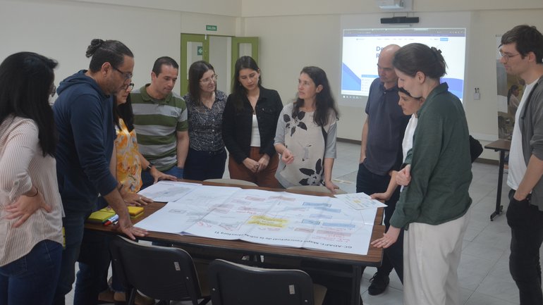 GC-ADAPT team members gathered around a table reviewing large printed diagrams during a workshop, with a presentation projected on the screen in the background.