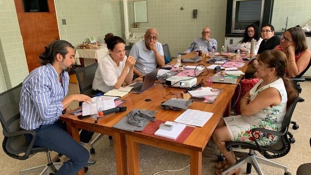 A group of international academic and public health stakeholders sitting together at a table, engaged in animated discussion. Paper materials and laptops are visible on the table.
