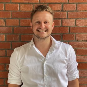 Yannick Daniel Höppner: a doctoral student stands outside in front of a brick wall and laughs into the camera.