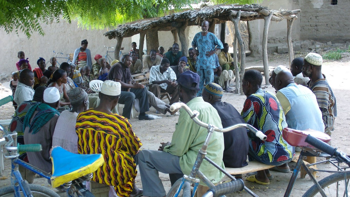 Community sensitisation in rural Tanzania Eine Gruppe sitzt auf einem Platz und hört einer Person zu, die spricht.