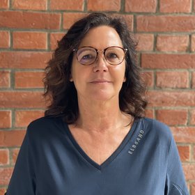 Heidrun von Thien: eine technische Assistentin mit mittellangen Haaren und braun umrandeter Brille vor einer Backsteinwand.