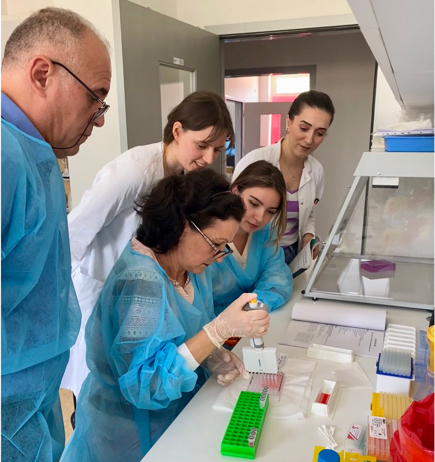 Labortraining beim Westbalkan Meeting 2025 Foto: Labortraining beim Westbalkan Meeting 2025. Mehrere Wissenschaftler:innen in blauen Laborkitteln beugen sich über eine Bench.