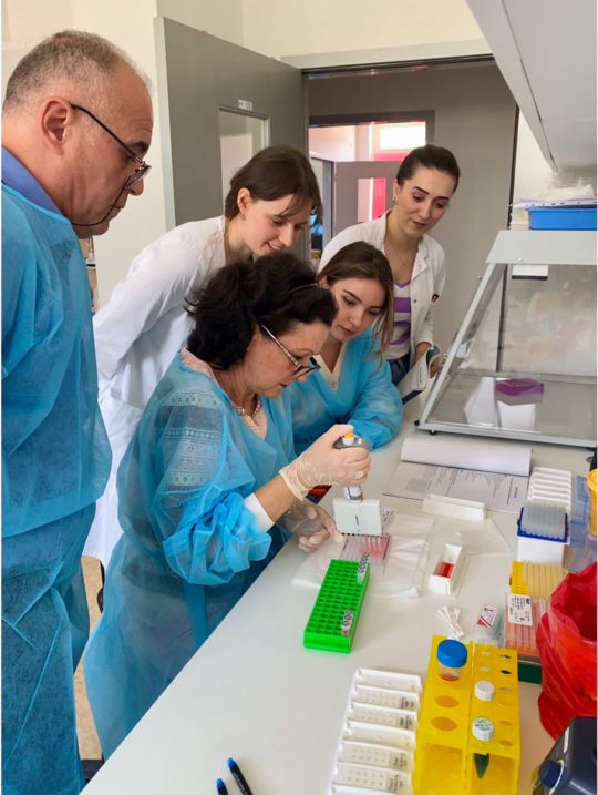 Labortraining beim Westbalkan Meeting 2025 Foto: Labortraining beim Westbalkan Meeting 2025. Mehrere Wissenschaftler:innen in blauen Laborkitteln beugen sich über eine Bench.