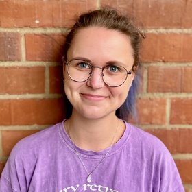 Janna Scherf: A PhD student smiles into the camera. She wears a pigtail, round glasses and a purple shirt.