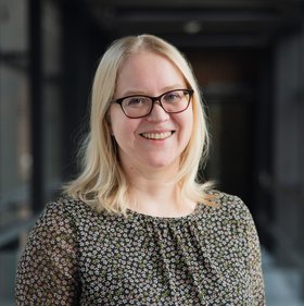 Foto von Anna Hein: eine Frau mit mittellangen, hellblonden Haaren, sie trägt ein dunkles Oberteil mit hellem Blümchenmuster.